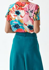 Krótka bluzka z krótkim rękawem w wielokolorowy wzór kwiatowy w odcieniach różu, czerwieni, niebieskiego i żółtego, zestawiona z turkusowymi spodniami. Gładka faktura tkaniny.