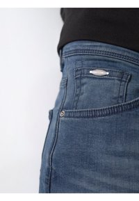 Jeans de mezclilla azul oscuro con cintura alta, que presentan un cierre frontal, botón de metal y una etiqueta de logo plateada en el bolsillo trasero. Textura sutil.