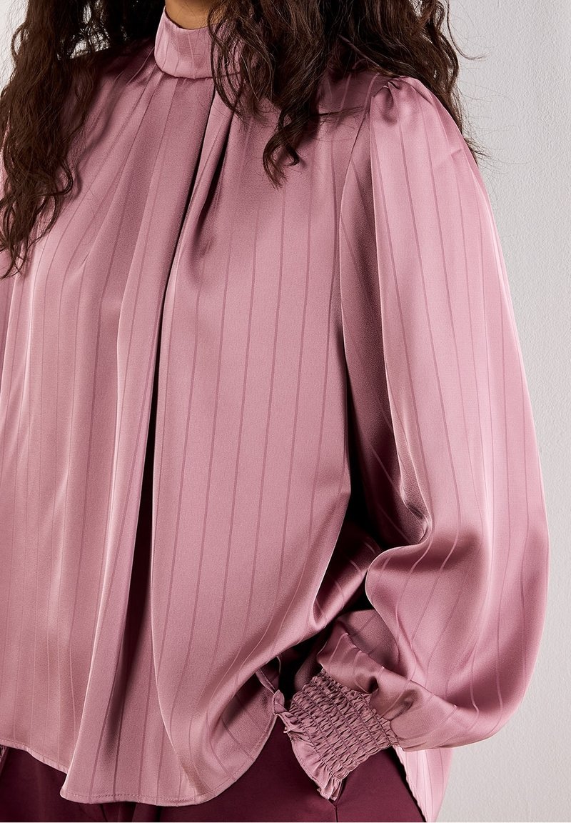 Femme portant un chemisier en satin mauve à manches longues, avec de subtiles rayures verticales et des poignets froncés, vue des épaules aux hanches.