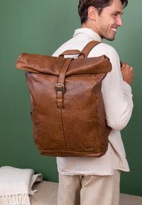 Sac à dos en cuir marron à rabat roulé avec des sangles réglables et quincaillerie en métal, doté d'une poche latérale pour un accès facile. Texture lisse et design minimal.