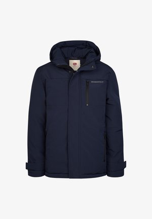 Dreng smilende iført en mørkeblå jakke, grønblå sweater, lyse revnede jeans og brune støvler, står med den ene hånd i lommen.