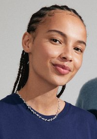 Sølv halskæde med et bølget design, båret over en marineblå t-shirt. Håret stylet i flettede sektioner for et markeret look.