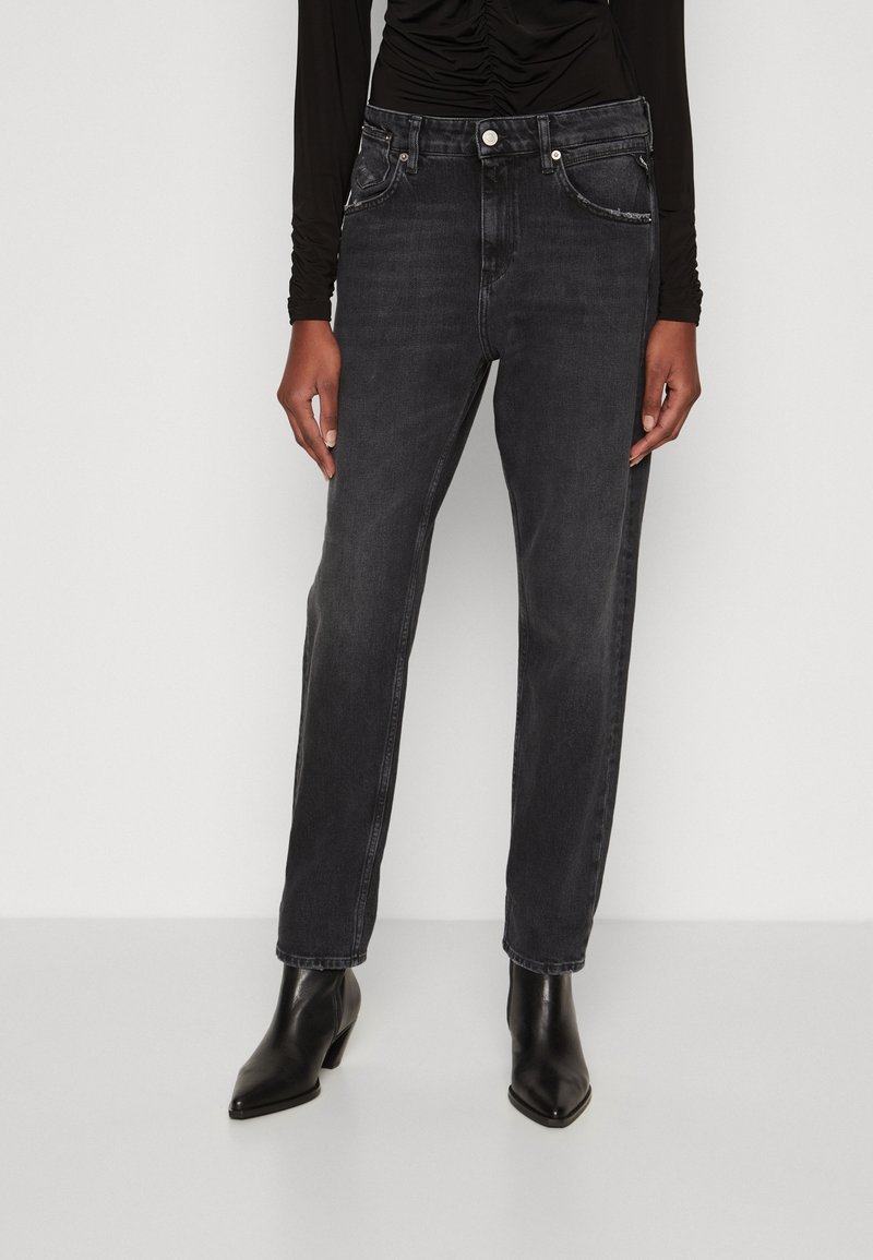 Personne portant un haut noir à manches longues, un jean noir coupe ample et des bottines noires, debout devant un fond uni clair.
