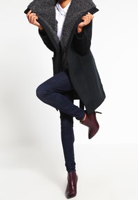 Cappotto nero con colletto foderato in pelliccia grigia, abbinato a jeans skinny blu scuro e stivali alla caviglia bordeaux, in piedi su una gamba.