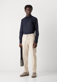 Camisa azul marino de botones, pantalones elásticos beige, bolso gris de hombro y zapatillas de ante gris claro. Diseño simple y ajustado con una paleta neutral.