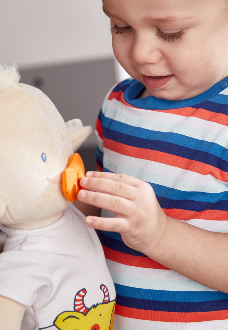 Plush toy with beige fabric, orange beak, and a white T-shirt featuring a cartoon monster with red and white horns; child touches the beak.
