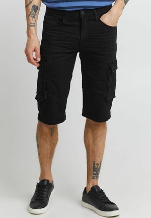 Mann trägt schwarze knielange Cargo-Shorts mit Taschen und schwarze Sneakers, steht mit einer Hand in der Tasche vor schlichtem Hintergrund.