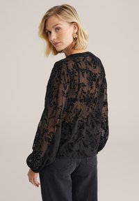 Blusa nera trasparente con ricami floreali, maniche lunghe a sbuffo, scollatura tonda e finitura testurizzata. Abbinata a jeans scuri.