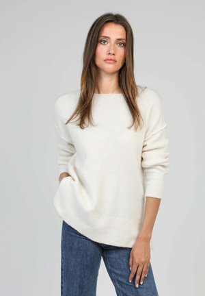 Femme aux cheveux longs et bruns portant un pull blanc à manches longues et un jean bleu, debout devant un fond gris uni.