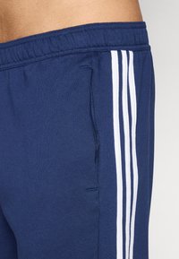 Pantalones deportivos azul marino con rayas verticales blancas. Tejido suave, cinturilla elástica, bolsillo lateral y una textura lisa.