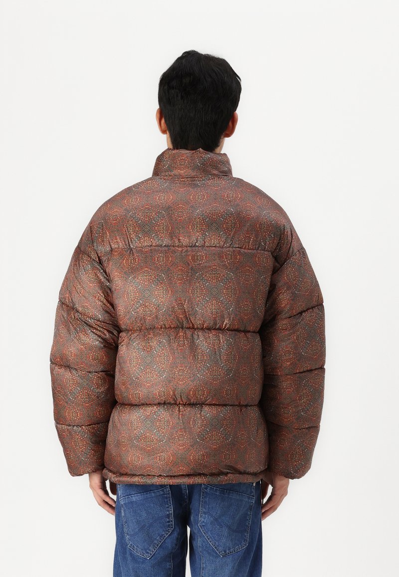 Braune gemusterte Daunenjacke mit hohem Kragen, kuscheliger Textur und auffälligen, gepolsterten Abschnitten; getragen über blauen Jeans.