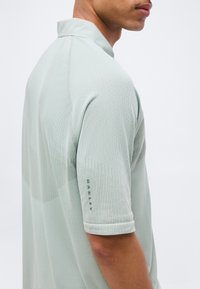 Άνδρας που φοράει ανοιχτοπράσινο, υφασμάτινο κοντομάνικο πουκάμισο, εμφανιζόμενος από πλάγια και πίσω, με φόντο μια απλή επιφάνεια.