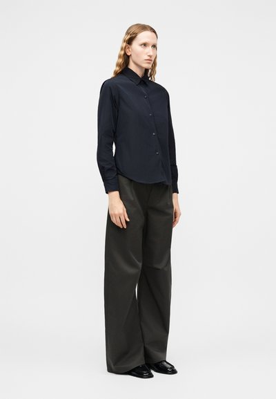 Femme debout portant une chemise noire à manches longues boutonnée, un pantalon large noir et des chaussures noires, sur un fond clair uni.