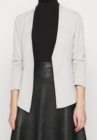 Jasnoszara marynarka z otwartym przodem, o długości rękawów do łokcia, miękka w dotyku z delikatnym marszczeniem, zestawiona z czarnym golfem i skórzaną spódnicą.