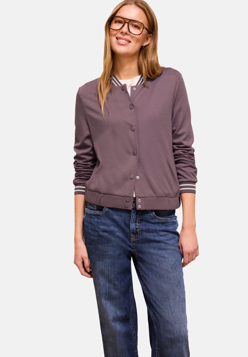 Donna sorridente che indossa occhiali grandi, una giacca bomber color malva con bottoni e polsini a righe, e jeans di denim blu, in piedi contro uno sfondo semplice.