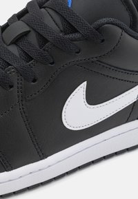 Gros plan d'une chaussure de sport noire avec des lacets noirs, le logo Nike Swoosh blanc et une semelle blanche montrant des coutures et une texture détaillées.