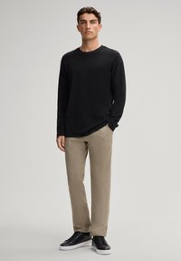 Langärmliges schwarzes T-Shirt kombiniert mit beigen Hosen. Das Modell trägt schwarze Sneakers. Schlichtes Design, neutrale Farben und lockere Passform.