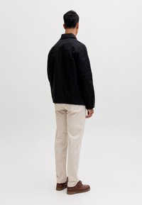 Bărbat stând cu fața spre depărtare, purtând o jachetă neagră, pantaloni bej și pantofi maro pe un fundal simplu și deschis.