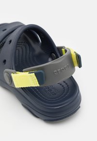 Laivastonsininen sandaali harmaalla säädettävällä nauhalla ja keltaisella korostuksella. Valmistettu kumista, jossa on tuuletusaukot ja teksturoitu pohja pitoa varten.