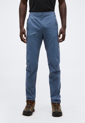 Mann trägt blaue Outdoor-Hose mit Reißverschlusstasche am Oberschenkel und braunen Wanderschuhen, steht vor einem einfarbigen Hintergrund.