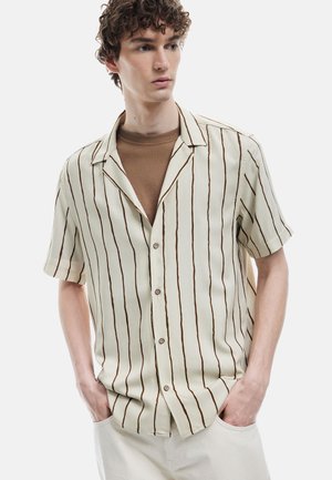 Camisa de manga corta en crema con rayas verticales marrones. Presenta un cuello tipo notch y cierre de botones, combinada con un suéter de cuello redondo marrón.