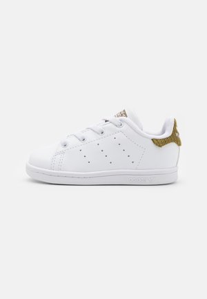 Baskets blanches Adidas Stan Smith avec lacets blancs, bandes perforées et languette de talon marron texturée sur un fond blanc uni.