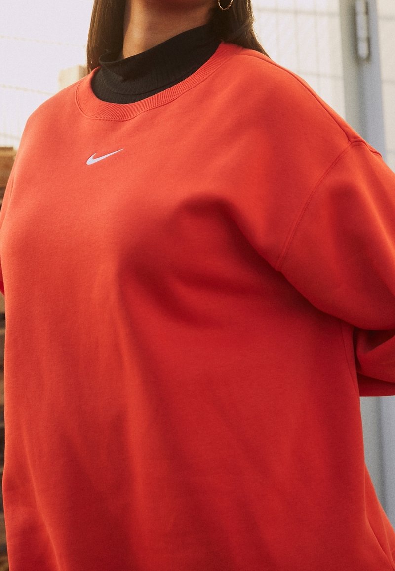 Röd sweatshirt i mjukt tyg med en ribbad krage och en liten vit Nike-logotyp på bröstet. Hänger något av axeln.