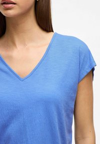 Niebieska koszulka z dekoltem w serek z krótkimi, podwiniętymi rękawami. Materiał wydaje się miękki i teksturowany, z widocznym szwem wzdłuż dekoltu.
