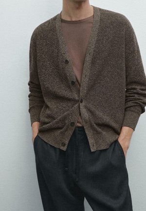 Mann trägt eine braune, geknöpfte Strickjacke über einem braunen T-Shirt, mit den Händen in den Taschen einer lockeren schwarzen Hose vor einem einfarbigen hellen Hintergrund.