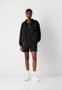 Svart hoodie med dragkedja och fickor, svart linne och svarta shorts. Vita sneakers och ankellånga strumpor fulländar outfiten.