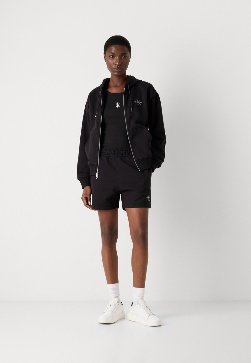 Zwarte hoodie met rits en zakken, zwarte tanktop en zwarte shorts. Witte sneakers en enkelsokken maken de outfit compleet.