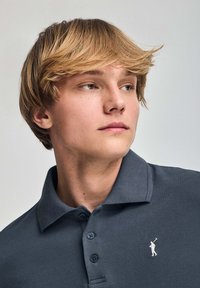 Polo en tela gris oscuro que presenta un cuello, tapeta de tres botones y un pequeño logotipo blanco de un golfista en el lado izquierdo del pecho.
