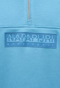 Κοντινό πλάνο μπλε υφάσματος με ασημένιο φερμουάρ και ανάγλυφη ορθογώνια ετικέτα λογότυπου που γράφει "NAPAPIJRI geographic."