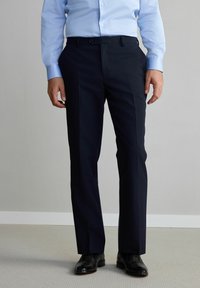 Pantalons de costume bleu marine en tissu lisse, présentant un design à devant plat, des jambes droites et des poches latérales. Complétés par des chaussures de ville noires.