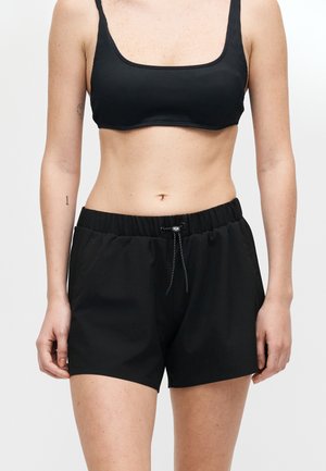 Femeie care poartă un sutien sport negru și pantaloni scurți negri cu talie elastică și șnur, pe un fundal simplu, deschis.