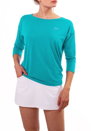 Nainen, jolla on päällään turkoosi pitkähihaiseen paitaan, jossa on pieni logo rinnassa, ja valkoinen hame, seisoo käsi taskussa.