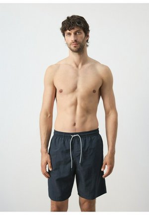 Hombre sin camiseta con cabello rizado castaño, vistiendo pantalones cortos de baño negros con cordón blanco, de pie contra un fondo liso y claro.