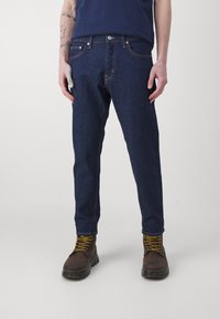 Slim-fit mørkeblå denimjeans med orange søm og fem lommer, kombinert med brune snørestøvler med gule lisser.