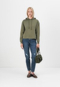 Khaki kapucnis pulóver zsinórokkal, kopott kék farmer, és fekete-fehér csíkos lapos cipők. A modell egy zöld, textúrált táskával áll.