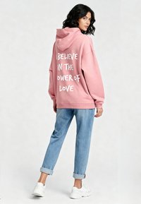 Felpa oversize rosa con la scritta bianca "CREDO NEL POTERE DELL'AMORE," abbinata a jeans azzurri risvoltati e sneakers bianche.