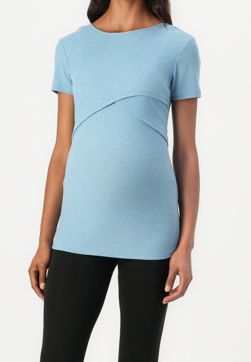Femme portant un haut de maternité à manches courtes côtelé bleu clair avec des leggings noirs, montrée du cou aux hanches sur fond blanc.