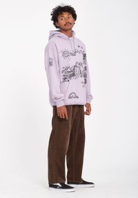 Haut à capuche lavande avec des impressions graphiques, dotée d'une poche kangourou et de côtes sur les côtés, accompagnée d'un pantalon en velours côtelé marron et de chaussures noires sans lacets.