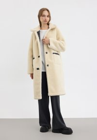 Cappotto in pile color crema con cappuccio, chiusure frontali a toggle, dettagli neri a contrasto sulle tasche e vestibilità ampia, abbinato a pantaloni scuri.