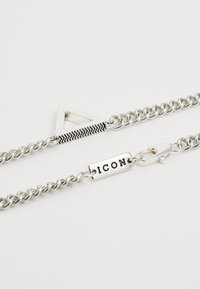 Bracelet en chaîne argentée avec un charm triangular, un bandeau noir texturé et une étiquette rectangulaire gravée "ICON."