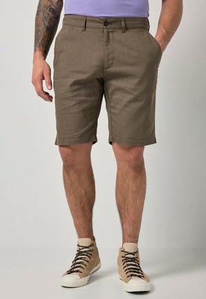 Mand iført brune knælange shorts, beige høje sneakers og en lilla skjorte, stående med den ene hånd i lommen.
