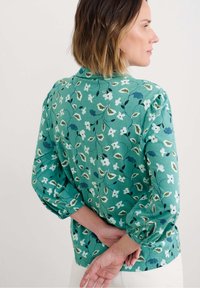 Grüne Bluse mit Puffärmeln, die mit weißen und dunkelblauen Blumenmustern verziert ist, abgeschlossen mit einem geraden Saum und weicher Textur.
