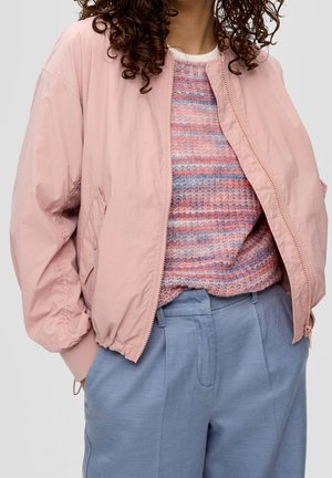 Lockighaarige Person trägt eine pinke Bomberjacke, einen mehrfarbig gestreiften Pullover und hellblaue hochtaillierte Hose mit Händen in den Taschen.