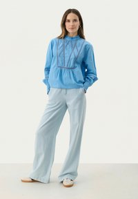 Blusa di cotone azzurro chiaro con dettagli ricamati elaborati, abbinata a pantaloni palazzo azzurro chiaro e sneaker bianche.