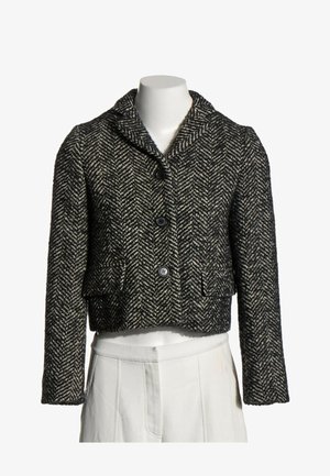Chaqueta de lana con patrón de espiga en blanco y negro, con tres botones y bolsillos delanteros con solapa en un torso de maniquí blanco.
