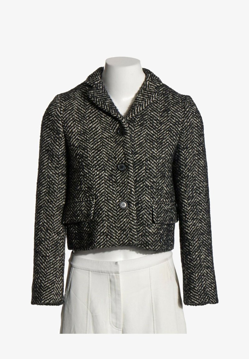 Veste en laine à motif chevrons noir et blanc avec trois boutons et poches à rabat avant sur un buste de mannequin blanc.